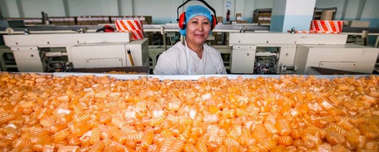 A woman machine operator smiles at an assembly line in Bayan-Sulu modern confectionery factory. Photo credit: Shutterstock.