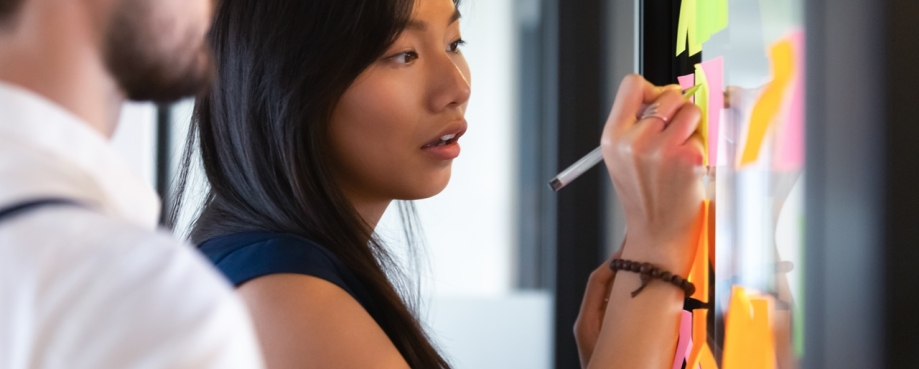 Focused woman writing an idea on post it sticky note on glass wall during a training workshop. Photo credit: Shutterstock.