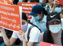 Masked women sit in protest against the military coup, holding red signs in English and Burmese. Photo credit: Wikicommons/ MgHla.