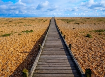 A long straight wooden decking pathway with a diminishing perspective on a pebble beach, the pathway starts large in the foreground centre of the image and becomes smaller as it reaches the horizon. Photo credit: Shutterstock. 