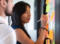Focused woman writing an idea on post it sticky note on glass wall during a training workshop. Photo credit: Shutterstock.